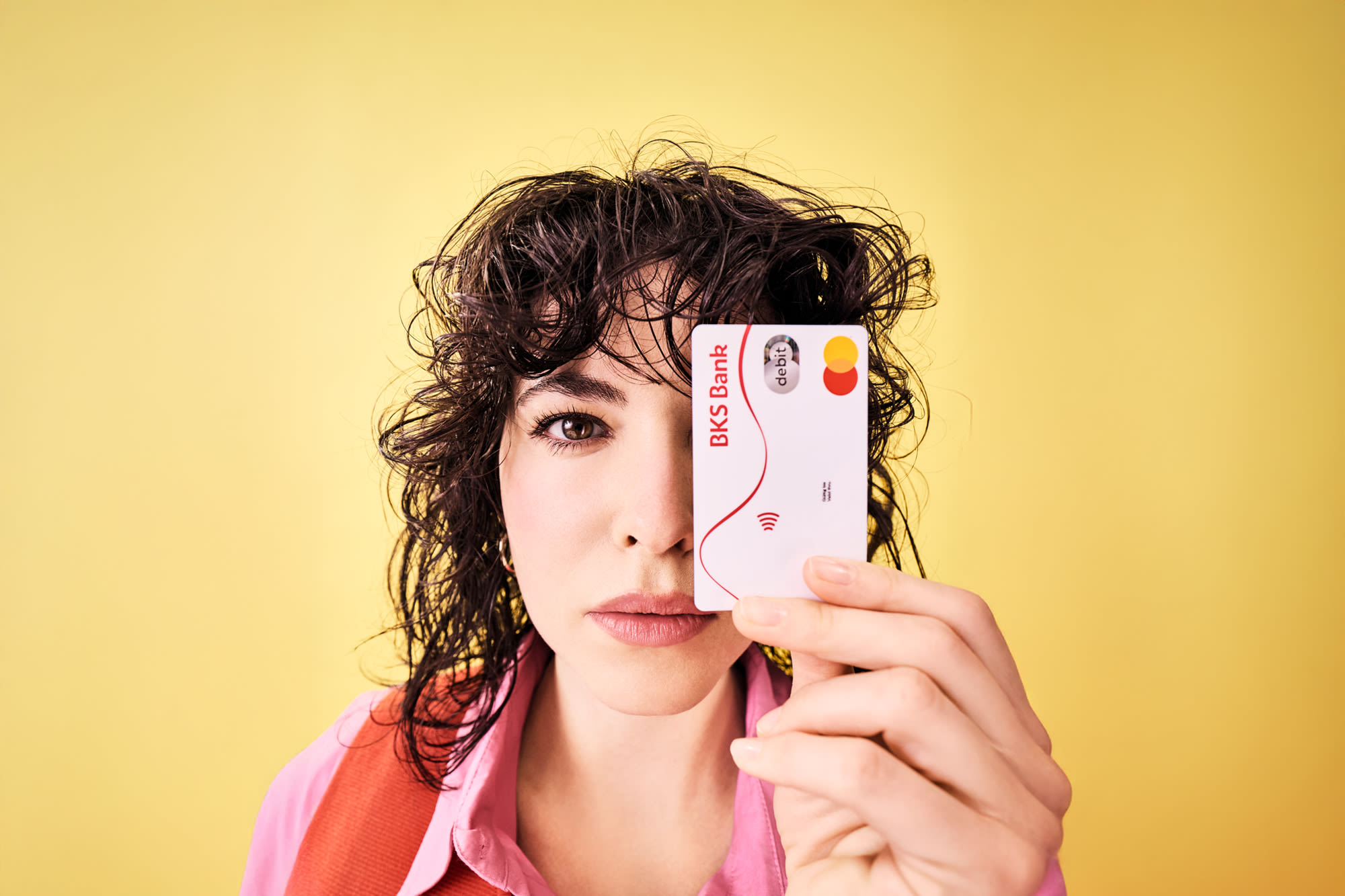 Ženska drži debetno kartico BKS neposredno pred obrazom in stoji pred rumenim ozadjem.