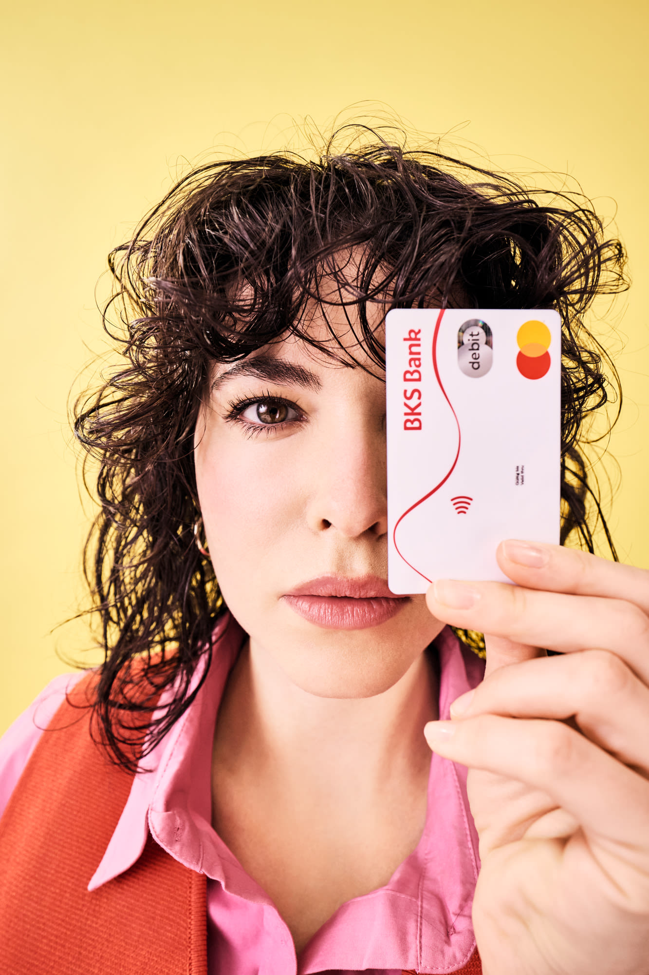 Ženska drži debetno kartico BKS neposredno pred obrazom in stoji pred rumenim ozadjem.